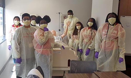 A gropu of children in masks and hospital gowns standing around a hostpital bed with a medical dummy laying on it.