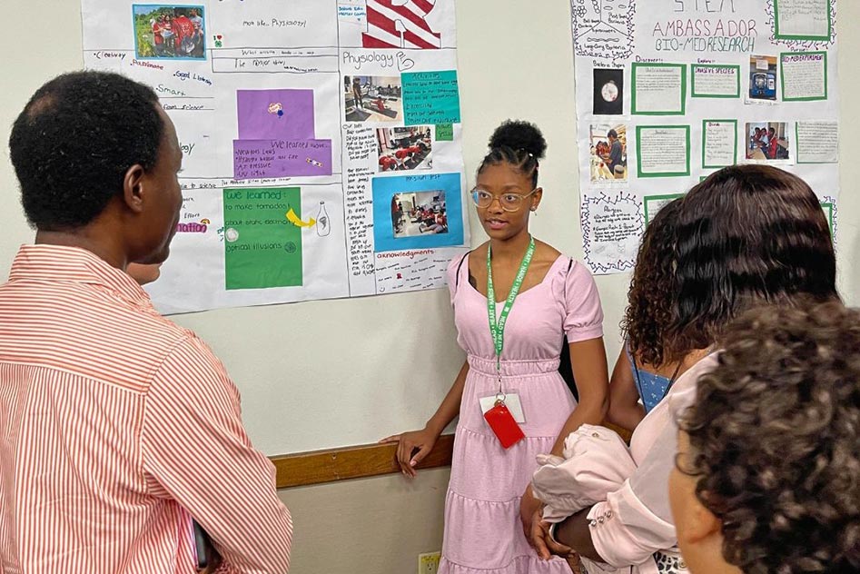 A returning STEM Ambassador presenting her final reflection presentation to family and friends. Behind her is her presentation poster, titled 'S. Technology. E. M. more like...physiology', reflecting her time working with an exercise scientist during this training.