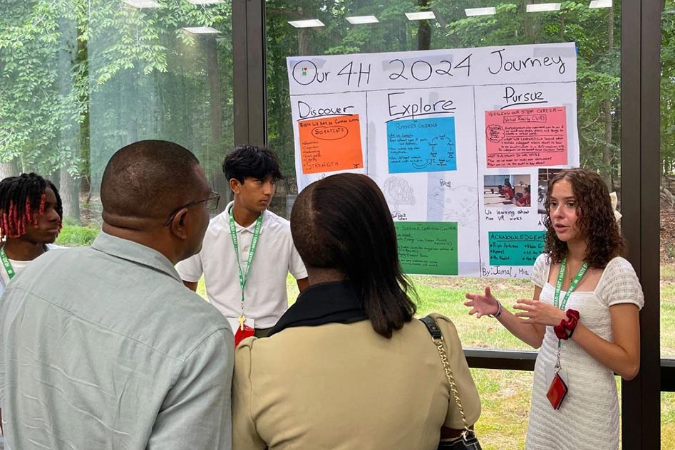 A returning STEM ambassador watching on as his presentation partner, a future STEM ambassador, presents on their final reflection poster to two adults. Their poster is titled 'Our 4H 2024 Journey' and features their time working with virtual reality and gaming specialists.