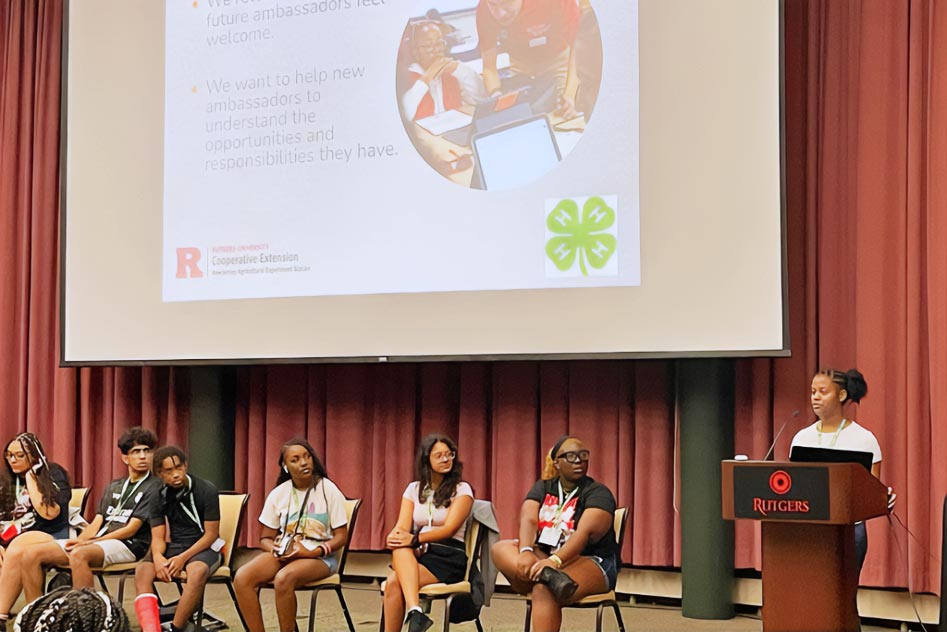 Six returning STEM Ambassadors sitting in front of a presentation as a panel. Above them is a projected slide titled 'Why We Came Back'. The sat ambassadors have their heads turned, listening to a peer and returning STEM Ambassador standing and talking at a Rutgers podium next to them. She is explaining that people return to make future ambassadors feel welcome and help them understand the opportunities and responsibilities they have. Those words are also on the projected slide.