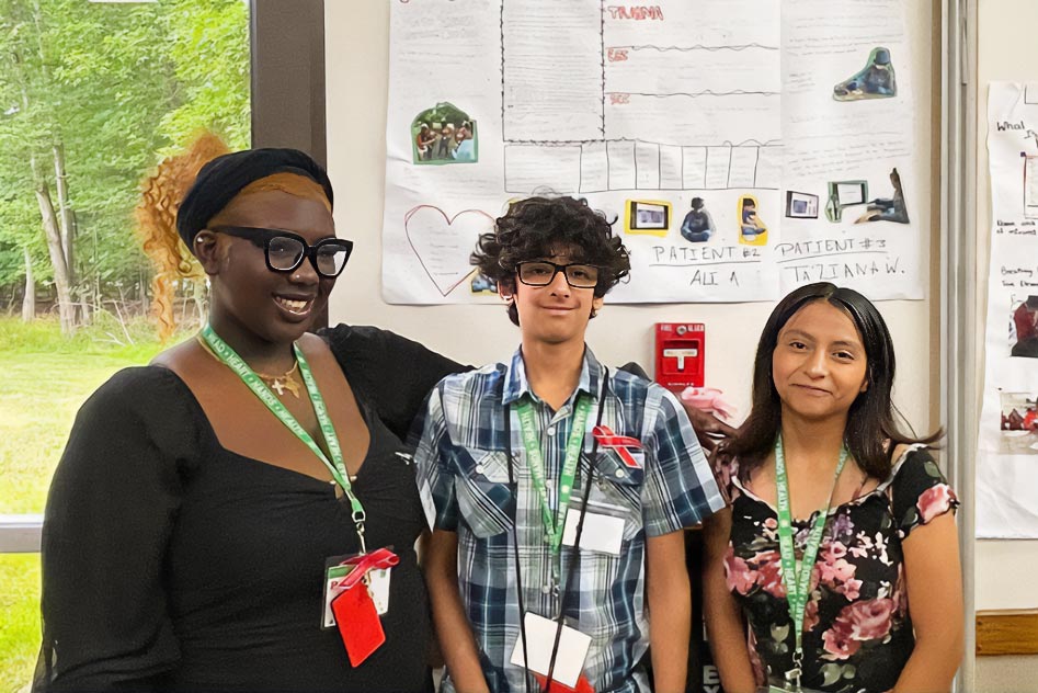 A returning STEM ambassador posing with two future STEM ambassadors. They worked together to create a final reflection and presentation on their time working with a doctor during this training. Behind them is a poster titled 'Acute Care Surgery'. The poster has 'Explore' and 'Discover' sections detailing their experiences.