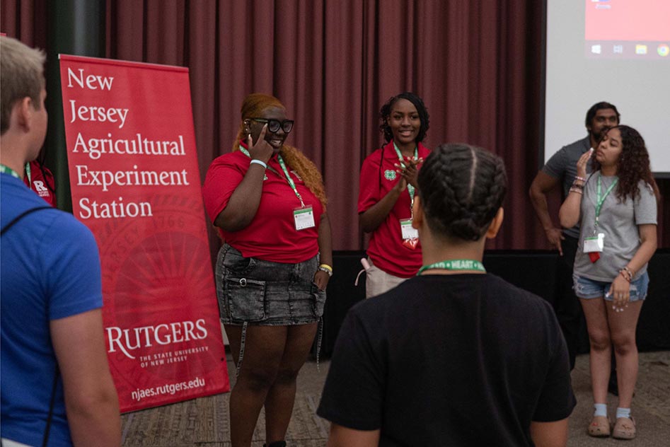 Two returning STEM Ambassadors leading a group icebreaker with the future STEM ambassadors. Behind them is a banner advertising the New Jersey Agricultural Experiment Station at Rutgers.