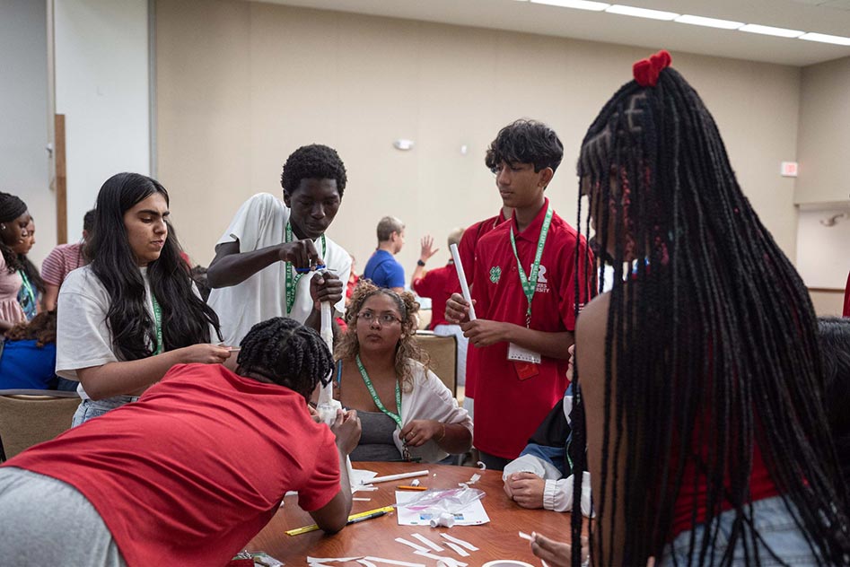 A returning STEM ambassador watches on as his group of future STEM ambassadors tackle an engineering challenge–balancing a can of beans on a paper tower. The five future ambassadors are experimenting with the height and shape of the tower. One of them is stabilizing the tower at its base. Another is shaping the top of the tower.